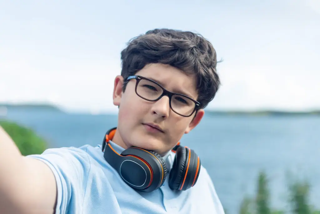 Portrait of eleven year old boy in glasses and wireless headphones, taking a selfie in front of the sea